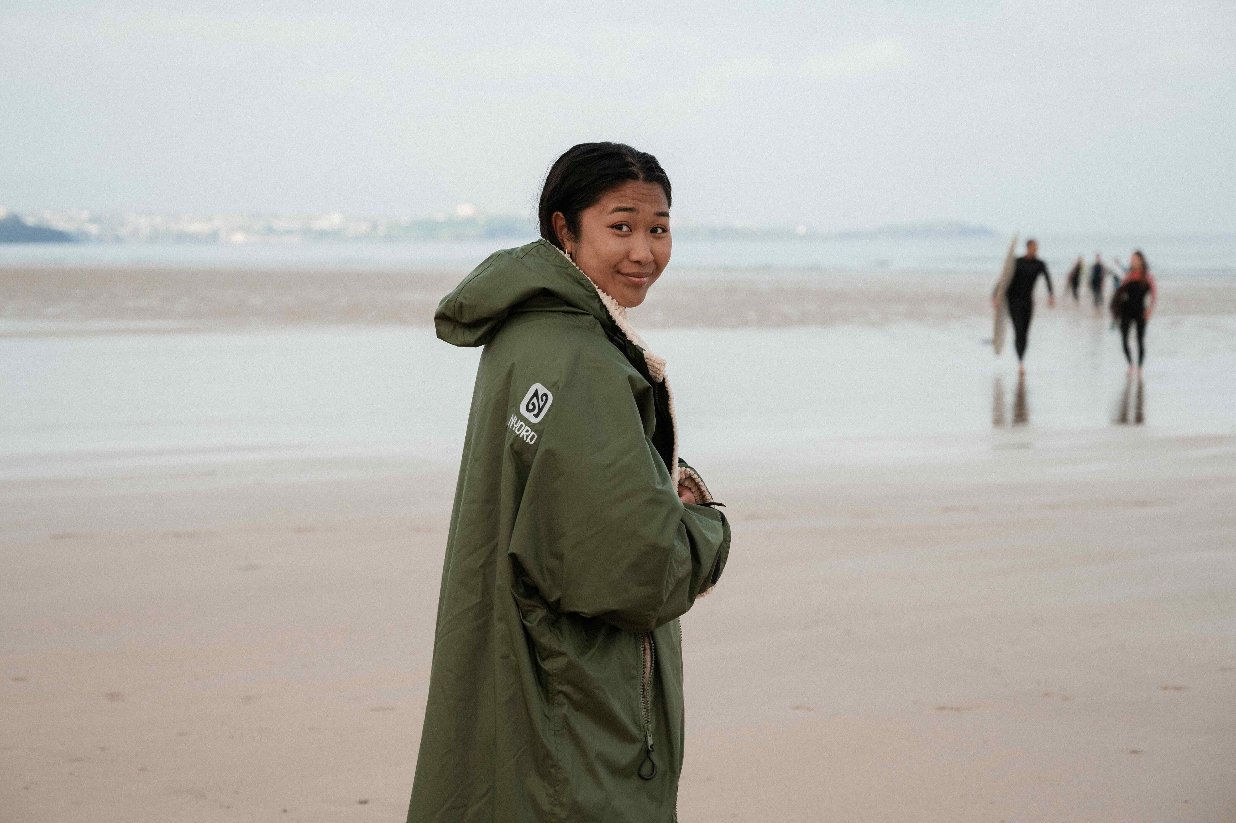 Une femme regarde par-dessus son épaule sur une grande plage, en poncho de change vert olive à capuche