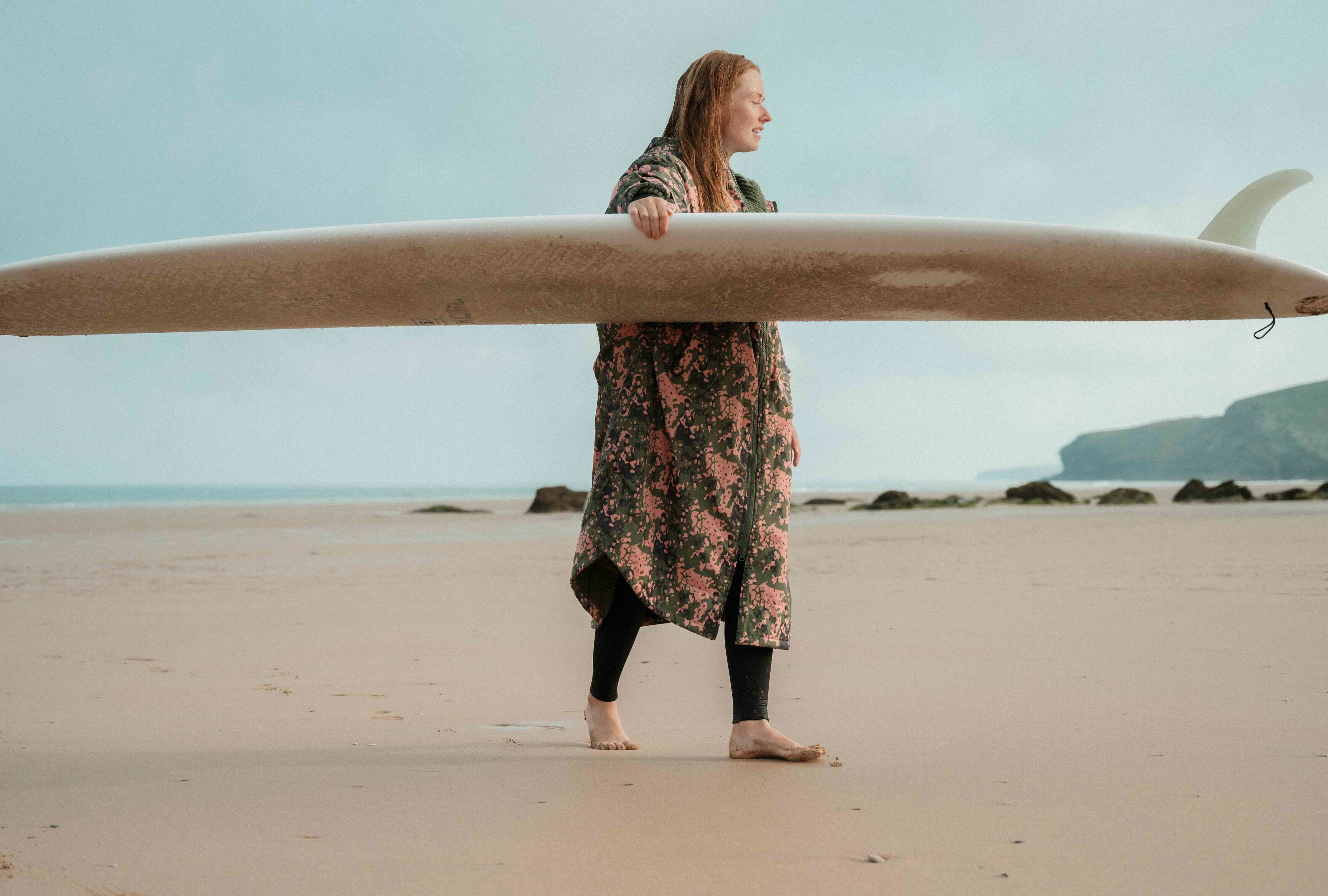 Une femme en robe à fleurs porte un longboard sur la tête le long du sable
