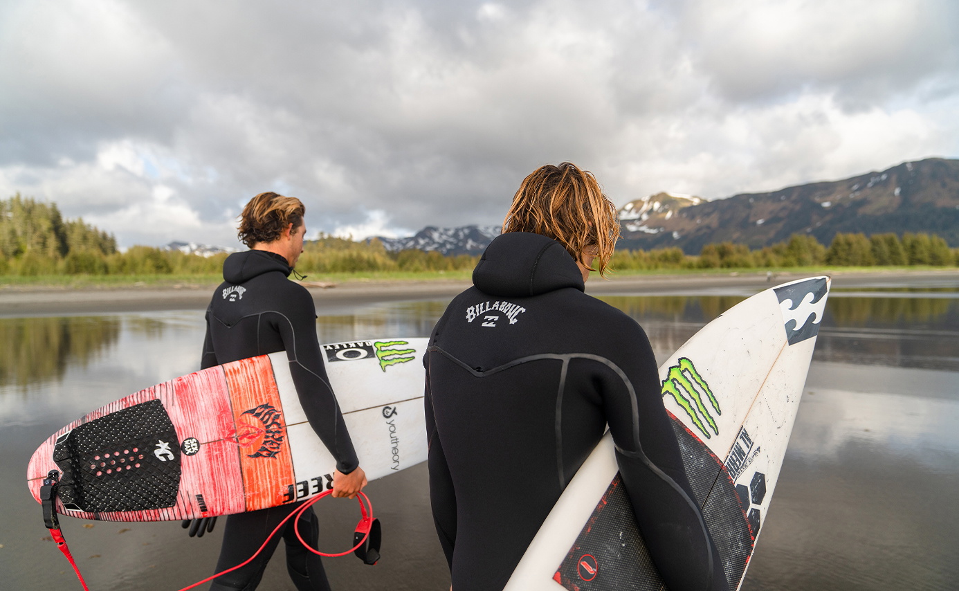 Two surfers in black wetsuits carry a surfboard and bodyboard along a shore backed by mountains
